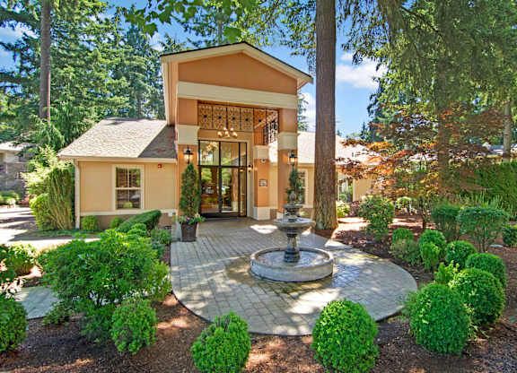 A welcoming leasing office entrance here at Asbury Park showcasing a charming building with a covered entry, glass front doors, and warm exterior accents, centered around a decorative fountain on a paved walkway, surrounded by manicured shrubs, mature trees, and landscaped greenery that create an inviting, park-like community setting.