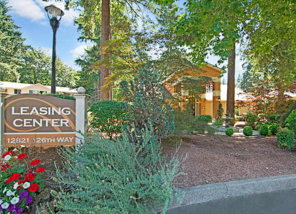 A landscaped leasing center entrance here at Asbury Park featuring a prominent leasing center sign along a curved driveway, surrounded by colorful flowers, manicured shrubs, and mature trees, with the leasing office building visible in the background, warm exterior lighting, and a tranquil fountain creating a welcoming, garden-style community atmosphere.