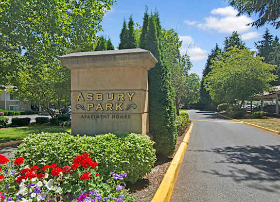 A landscaped community entrance sign here at Asbury Park featuring a stone monument displaying the property name, surrounded by colorful flowers, trimmed hedges, and mature trees, with a paved driveway extending into the community and covered parking visible in the background under a clear blue sky.
