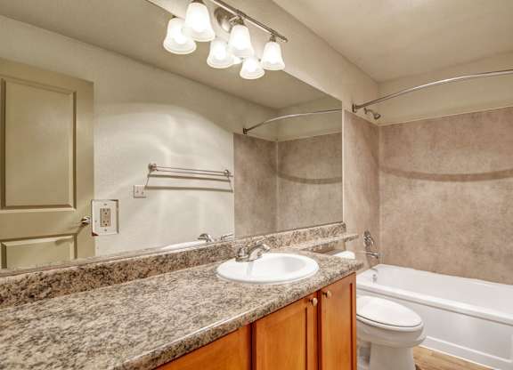A well-appointed bathroom here at Asbury Park showcasing a spacious granite-style vanity countertop with an undermount sink, warm wood cabinetry, and a wide wall mirror beneath bright overhead lighting, paired with a tub and shower combination featuring neutral wall surrounds, a curved shower rod, and coordinated fixtures for a clean, practical layout.