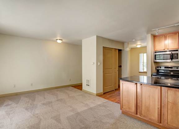 A spacious open living area here at Asbury Park featuring soft carpeted flooring, neutral walls, and a wide layout that flows into a functional kitchen with warm wood cabinetry, a dark countertop, and modern appliances, while ceiling lighting and a nearby window provide even illumination and a comfortable, practical atmosphere.