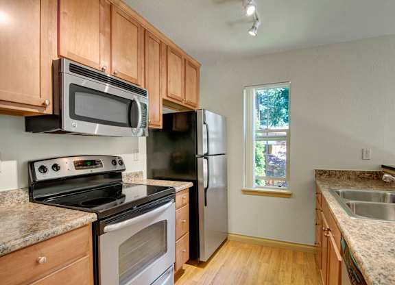 A functional galley kitchen here at Asbury Park featuring warm wood cabinetry, smooth countertops, and a coordinated appliance set with a built-in microwave, range, and refrigerator, complemented by wood-style flooring, a double-basin sink, and a window that brings in natural light for a clean, efficient cooking space.