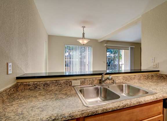 A welcoming kitchen pass-through here at Asbury Park showcasing smooth, speckled countertops with a double-basin sink and chrome faucet, opening to a dining and living area with vertical blinds, a sliding glass door, and a pendant light that adds warmth while allowing natural light to flow through the connected space.