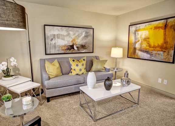 A cozy living room here at Colonial Square featuring a soft gray sofa with yellow and patterned accent pillows, a sleek white coffee table with decorative vases, and a side table with a modern lamp, complemented by abstract wall art, neutral carpeting, and warm ambient lighting that creates an inviting and comfortable atmosphere.