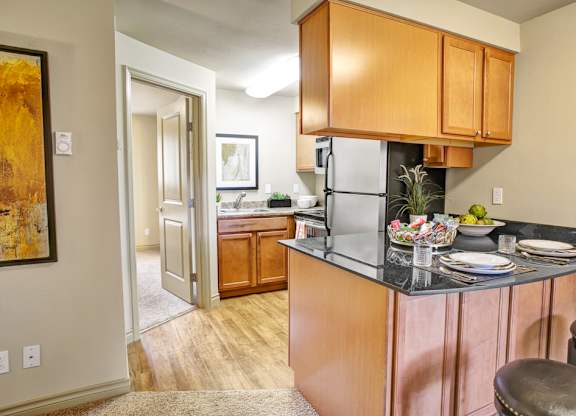 A compact kitchen here at Colonial Square featuring warm wood cabinetry, dark countertops with bar seating, and a small breakfast bar set with plates and glassware, complemented by wood-style flooring, a stainless-look refrigerator, a nearby sink area with additional cabinets, and soft overhead lighting that creates a functional and welcoming cooking space.