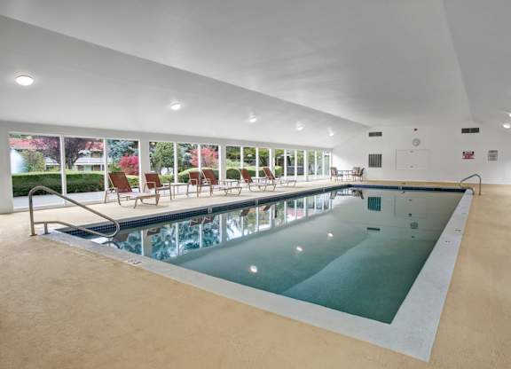 A spacious indoor swimming pool here at Colonial Square featuring a long rectangular pool with stainless steel handrails, a wide pool deck with lounge chairs arranged along floor-to-ceiling windows, smooth light-toned flooring, recessed ceiling lighting, and views of landscaped greenery outside that create a calm and inviting atmosphere.