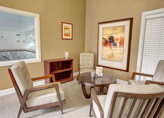 A welcoming lounge area here at Colonial Square featuring four cushioned armchairs with wood frames arranged around a round table, neutral carpeted flooring, framed artwork on warm-toned walls, and a small shelving unit, with large interior and exterior windows that add natural light and create a comfortable space for conversation and relaxation.