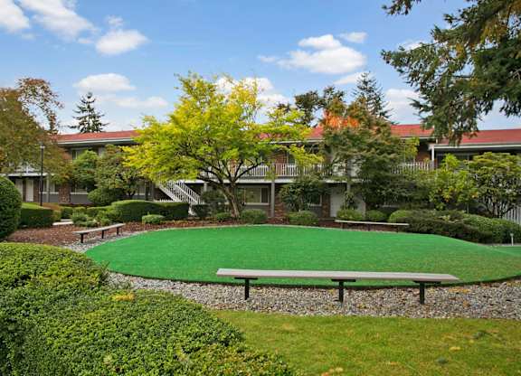 A landscaped courtyard lawn here at Colonial Square featuring a large circular green space bordered by gravel pathways and simple bench seating, surrounded by mature trees with seasonal foliage, neatly trimmed shrubs, and two-story apartment buildings with exterior walkways, creating a calm, park-like community gathering area.