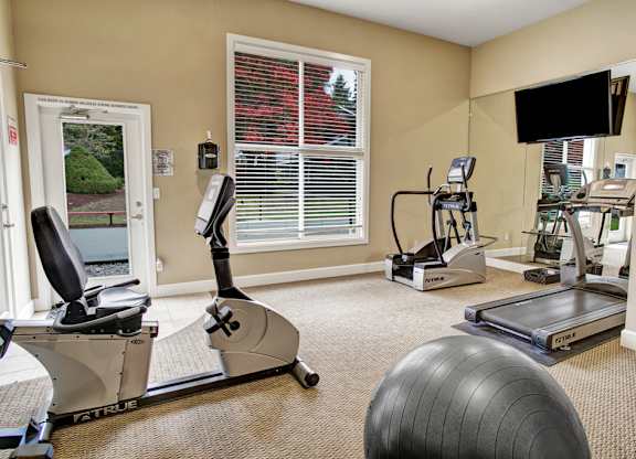 A compact fitness room here at Colonial Square featuring cardio equipment including a treadmill, stationary bike, and elliptical machine, complemented by a wall mirror, mounted television, large window with blinds bringing in natural light, and neutral carpeting that creates a clean and functional workout space.