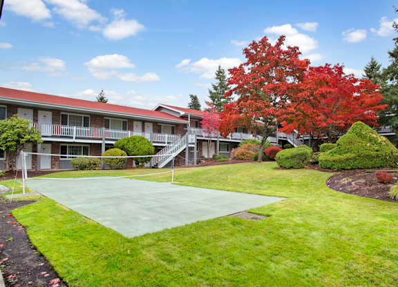A landscaped outdoor volleyball court here at Colonial Square featuring a smooth paved playing surface with a central net, bordered by lush green lawn, manicured shrubs, and mature trees with vivid autumn foliage, while surrounding apartment buildings with balconies and walkways create a welcoming, park-like community setting.