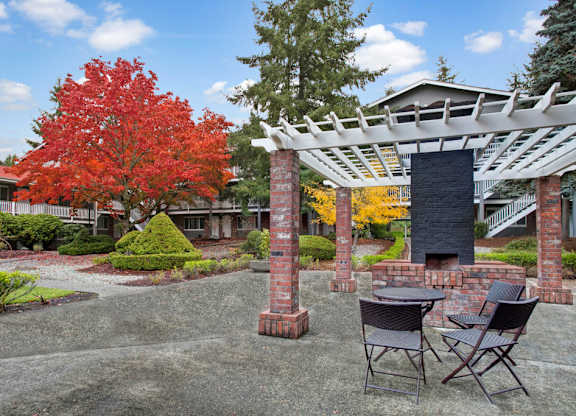 A landscaped outdoor courtyard here at Colonial Square featuring a brick pergola with a built-in grill, a small round table with chairs, and a paved seating area surrounded by manicured shrubs, gravel pathways, and vibrant red and yellow autumn trees, with two-story apartment buildings and exterior walkways in the background.