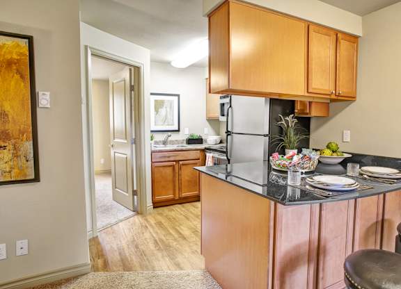 A compact kitchen here at Colonial Square featuring warm wood cabinetry, dark countertops with bar seating, and a small breakfast bar set with plates and glassware, complemented by wood-style flooring, a stainless-look refrigerator, a nearby sink area with additional cabinets, and soft overhead lighting that creates a functional and welcoming cooking space.