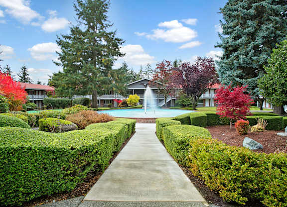 A landscaped courtyard here at Colonial Square featuring a central circular fountain with a tall water feature, surrounded by neatly trimmed hedges, colorful seasonal trees, and garden beds, with a straight concrete walkway leading toward the fountain and two-story apartment buildings with exterior walkways framing the peaceful, park-like setting.