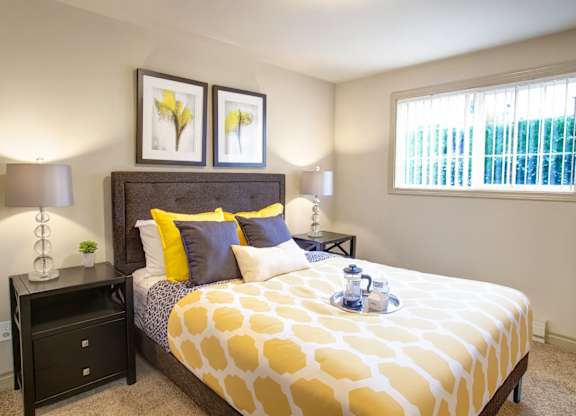 A cozy bedroom here at Colonial Square featuring a neatly made bed with a patterned yellow and white comforter, layered accent pillows, and a padded headboard, flanked by matching nightstands with modern lamps, complemented by framed wall art, soft carpeted flooring, and a window with vertical blinds providing natural light.