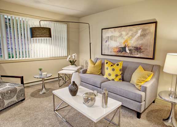 A cozy living room here at Colonial Square featuring a light gray sofa with yellow accent pillows, a white coffee table with decorative accents, soft carpeted flooring, and modern side tables with lamps, complemented by abstract wall art, a floor lamp, and a large window with vertical blinds that brings in natural light.