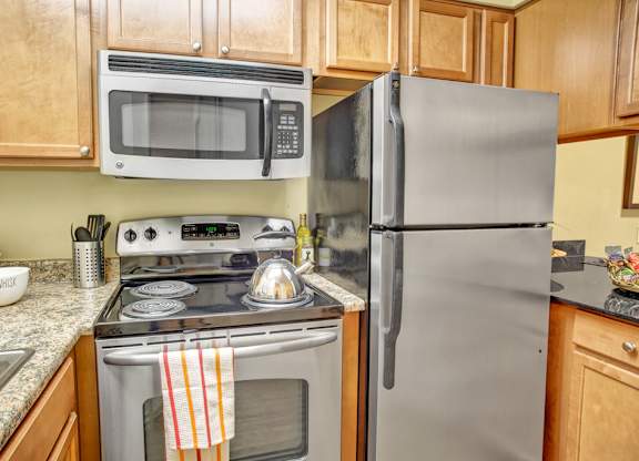 A compact kitchen here at Colonial Square featuring warm wood cabinetry, speckled countertops, a stainless refrigerator, a built-in microwave above a smooth cooktop range, and a sink set into the counter, with simple decor and neatly arranged cookware creating a functional, efficient space for everyday cooking and meal prep.