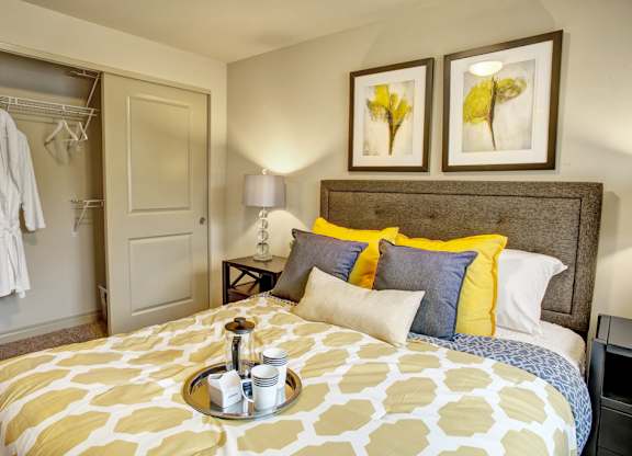 A welcoming bedroom here at Colonial Square featuring a neatly made bed with a patterned yellow and white comforter, layered accent pillows, and a textured upholstered headboard, accompanied by a nightstand with a table lamp, framed wall art above the bed, soft carpeted flooring, and an open closet with shelving and hanging space.