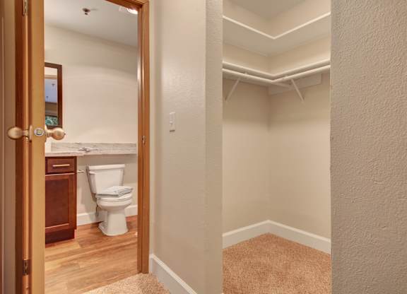 A spacious walk-in closet here at Forest Greens featuring beige carpet, neutral walls, and wraparound hanging rods with open shelving for storage. The closet sits just outside the bathroom, offering convenient access to a vanity area with a wood cabinet, light countertop, and toilet visible through the doorway.