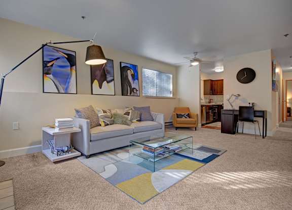 A stylish living room here at Forest Greens featuring a neutral sofa with patterned throw pillows, a glass coffee table over a modern area rug, and framed artwork displayed above the seating area. The space includes plush carpeting, a floor lamp, ceiling fan, and an open layout that flows toward a dining nook and kitchen in the background.