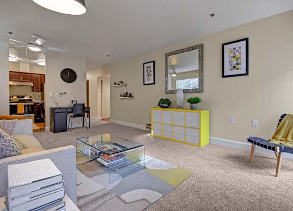 A contemporary living room here at Forest Greens featuring plush carpeting, a glass coffee table set over a patterned area rug, and a neutral sofa accented with decorative pillows. The space includes framed wall art, a modern console with greenery, a desk nook, and an open layout that connects to the kitchen and dining area.