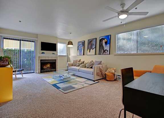 A stylish living room here at Forest Greens featuring plush carpet flooring, a neutral sofa with accent pillows, and a glass coffee table set on a patterned area rug. The space includes a cozy fireplace with a mounted TV above, large windows with blinds, sliding glass doors leading to a private patio or balcony, ceiling fan lighting, wall art, and a small dining or work desk area nearby.