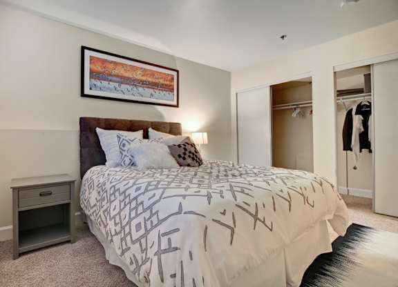 A comfortable bedroom here at Forest Greens featuring plush carpet flooring, a bed with patterned bedding and layered accent pillows, and a nightstand with a table lamp. The space includes a framed wall art piece above the headboard, sliding closet doors with open storage visible, neutral wall tones, and soft lighting that creates a warm, inviting atmosphere.