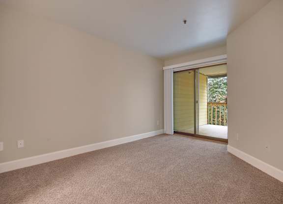 A spacious bedroom here at Forest Greens featuring soft carpet flooring, neutral wall tones, and an open layout that offers flexible furniture placement. The room includes a sliding glass door with vertical blinds that opens to a private balcony with a railing and leafy outdoor views, allowing natural light to brighten the space and create an airy, comfortable atmosphere.