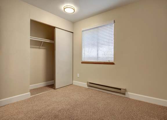 A cozy bedroom here at Forest Greens featuring soft carpet flooring, neutral wall tones, and a compact layout ideal for a guest room or home office. The space includes a sliding-door closet with open hanging storage, a window with white blinds that allows natural light, a baseboard heater below the window, and simple overhead lighting that keeps the room bright and functional.