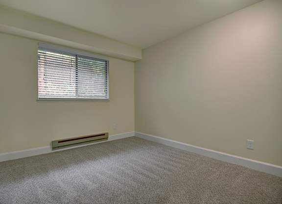 A bright bedroom here at Forest Greens featuring plush carpet flooring, soft neutral wall tones, and a simple open layout that allows for flexible furniture placement. The space includes a window with white blinds that brings in natural light, a baseboard heater below the window, clean white trim, and an uncluttered design that creates a calm, comfortable atmosphere.