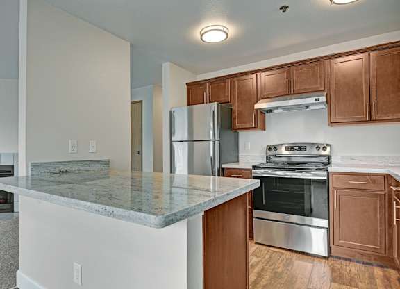 A spacious open kitchen here at Forest Greens featuring rich wood cabinets, light stone-style countertops, and a full appliance setup with a refrigerator, range, and hood. A wide breakfast bar overlooks the adjacent living area, while wood-look flooring and ceiling lights create a warm and functional cooking and gathering space.