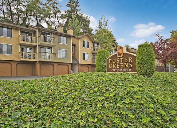 A welcoming exterior view here at Forest Greens showcasing the community monument sign set among manicured greenery, with multi-story apartment buildings in warm neutral tones behind it. The scene features private balconies, garage access below the residences, mature trees, and a landscaped setting that creates a peaceful residential atmosphere.