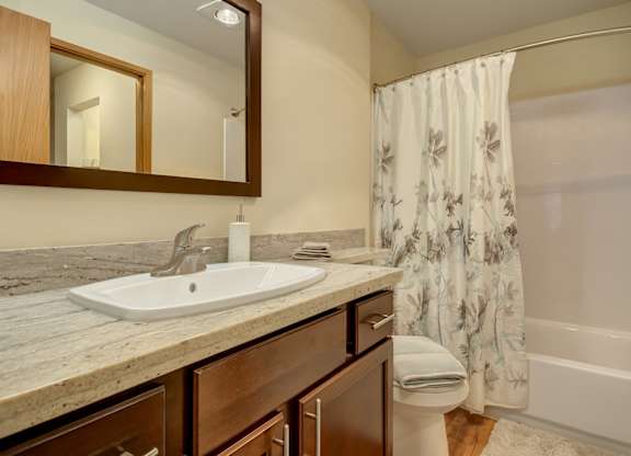 A clean bathroom here at Forest Greens featuring a wood vanity with a light stone-style countertop and integrated sink beneath a framed mirror. A bathtub-shower combination with white wall surround and patterned shower curtain sits beside the vanity, while wood-style flooring, folded towels, and neutral finishes create a tidy, welcoming space.
