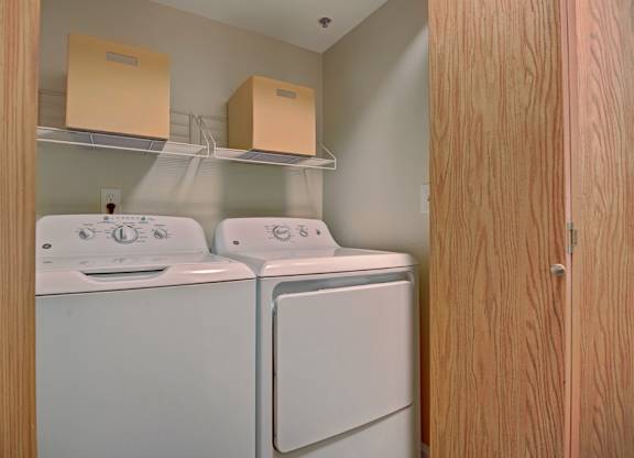 A convenient in-home laundry closet here at Forest Greens featuring a side-by-side washer and dryer tucked behind wood-paneled doors. White wire shelving above provides storage space for supplies, while neutral walls and a compact layout keep the area functional and easy to access.