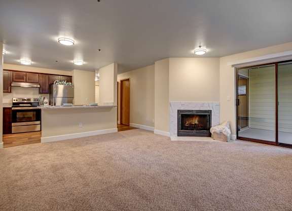 A wide open living room here at Forest Greens featuring plush beige carpet, soft neutral walls, and a tiled fireplace centered along one wall. The space opens to the kitchen with a breakfast bar and stainless steel appliances, while a sliding glass door leads to a covered patio, bringing in natural light and extending the living area outdoors.
