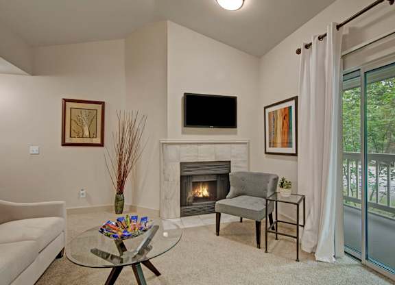 A cozy living room here at Island Park featuring soft neutral carpeting, a corner fireplace with a tiled surround, and a mounted television above the mantel. The space includes a comfortable accent chair, a glass coffee table with decorative accents, framed wall art, and sliding glass doors with curtains leading to a private balcony surrounded by greenery.