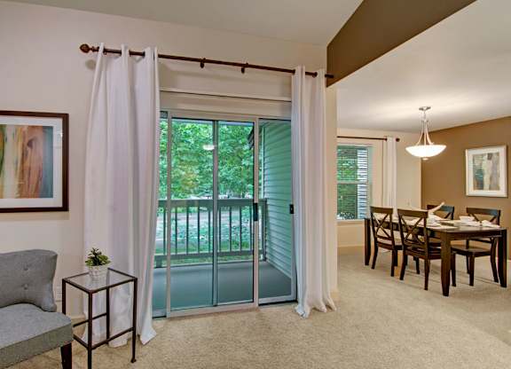 A comfortable living and dining area here at Island Park featuring soft carpeting, neutral walls, and a sliding glass door with white curtains that opens to a private balcony overlooking greenery. The living space includes an accent chair and side table, flowing into a dining area with a wood table, matching chairs, and a pendant light.