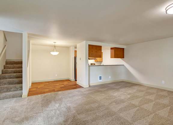 A spacious open living area here at Island Park featuring plush neutral carpeting, an open staircase with carpeted steps, and a dining nook with wood-style flooring and a hanging light fixture. The layout flows into a kitchen pass-through with upper cabinetry, creating an open, flexible space suitable for both living and dining.
