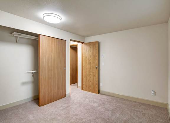 A simple unfurnished bedroom here at Island Park featuring soft neutral carpeting, an open closet with wood sliding doors and hanging shelves, and a doorway leading to the hallway. The room includes a large window with horizontal blinds allowing natural light to fill the space, offering a clean, flexible layout suitable for a bedroom or home office.
