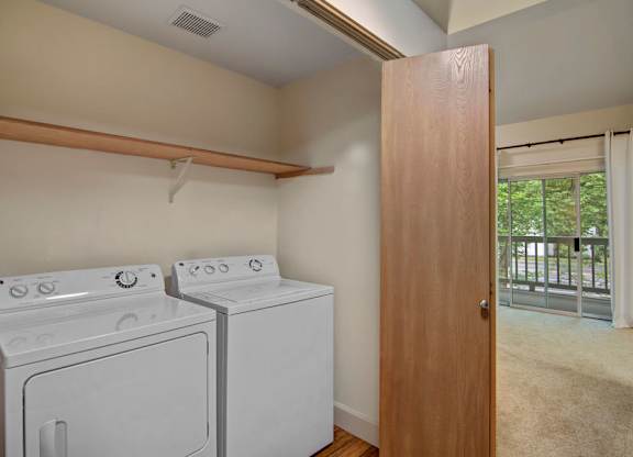 A convenient in-home laundry area here at Island Park featuring a full-size washer and dryer set beneath an open storage shelf, with wood-style flooring and neutral walls. The space opens to the living area through a wood door and sits near a sliding glass door that brings in natural light, creating a functional and easy-to-access layout.