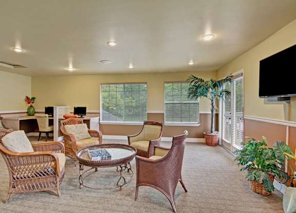 A welcoming community lounge here at Island Park featuring wicker seating arranged around a round coffee table, a mounted television, and multiple computer stations for resident use. Large windows and glass doors bring in natural light, while potted plants and neutral flooring create a comfortable, inviting space for relaxing or working.
