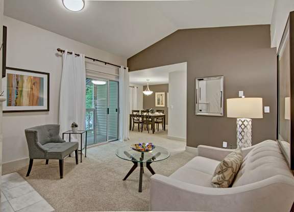 A spacious living room here at Island Park showcasing soft neutral carpeting, a comfortable sofa with patterned accent pillows, and a glass coffee table centered in the room. The space features an accent chair, framed wall art, a table lamp, and sliding glass doors with curtains that open to a private balcony, with an open view into the adjacent dining area.