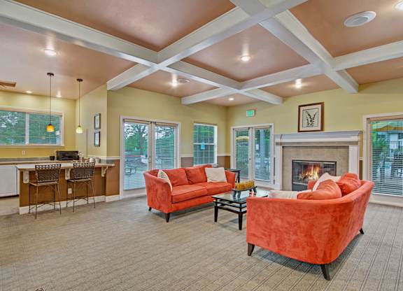 A welcoming community lounge here at Island Park featuring coffered ceilings, soft neutral flooring, and large windows that bring in natural light. The space includes plush accent sofas arranged around a fireplace, a glass coffee table, and a kitchenette with bar seating, creating a comfortable area for relaxing or gathering with neighbors.