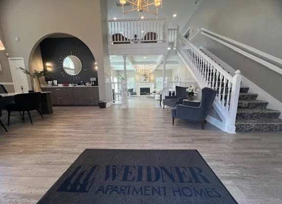 A bright and welcoming clubhouse lobby here at Island Park featuring wide plank flooring, a grand staircase with white railings, and an open two-story layout. The space includes comfortable seating areas, a reception desk, and elegant lighting that creates a warm first impression for residents and guests.