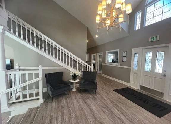A bright clubhouse entry here at Island Park featuring wide plank flooring, a white staircase with classic railings, and a comfortable seating area with two upholstered chairs and a small accent table. The space is accented by a modern chandelier, soft neutral walls, and glass-paneled doors that enhance the open, welcoming atmosphere.