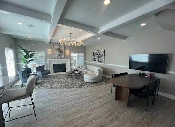 A spacious clubhouse lounge here at Island Park featuring wide plank flooring, a coffered ceiling with recessed lighting, and a cozy seating area arranged around a fireplace. The room includes a curved sofa, accent chairs, wall art, a mounted television, and a round table with chairs, creating a welcoming space for relaxing or gathering.