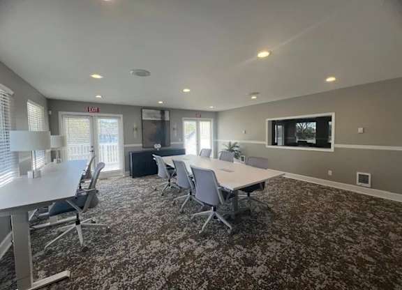 A modern meeting room here at Island Park featuring long conference tables with rolling chairs, patterned carpet flooring, recessed ceiling lighting, and neutral gray walls. The space includes large windows, glass double doors, a wall-mounted display, and a clean open layout designed for resident gatherings, meetings, or collaborative use.