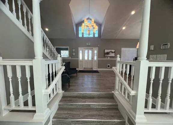 A welcoming community lobby here at Island Park featuring a central staircase with white railings, wood-look flooring, and a high vaulted ceiling with a decorative chandelier. The space includes neutral walls, comfortable seating areas, framed wall art, and glass entry doors that create an open, inviting atmosphere for residents and guests.
