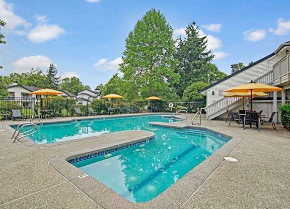 A refreshing outdoor swimming pool here at Island Park featuring a spacious concrete deck, a separate shallow pool area, and multiple lounge chairs with bright umbrellas. The fenced pool area is surrounded by mature trees, landscaped greenery, and nearby community buildings, creating a relaxing setting for residents.