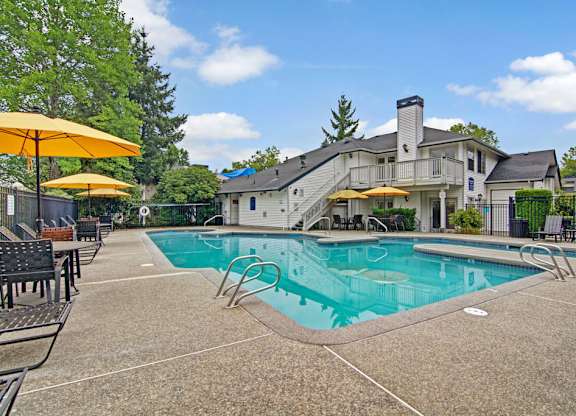 A resort-style outdoor swimming pool here at Island Park featuring a large rectangular pool with metal handrails, a wide concrete deck, and several lounge chairs shaded by bright yellow umbrellas. The fenced pool area is surrounded by mature trees, landscaped greenery, and nearby residential buildings under a blue sky.