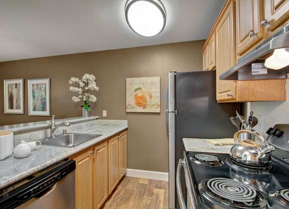 A functional kitchen here at Island Park featuring warm wood cabinetry, granite-style countertops, and a stainless-steel sink with a modern faucet. The space includes a full appliance suite, wood-look flooring, and coordinated wall art above the counter, creating a clean and practical layout with a welcoming feel.